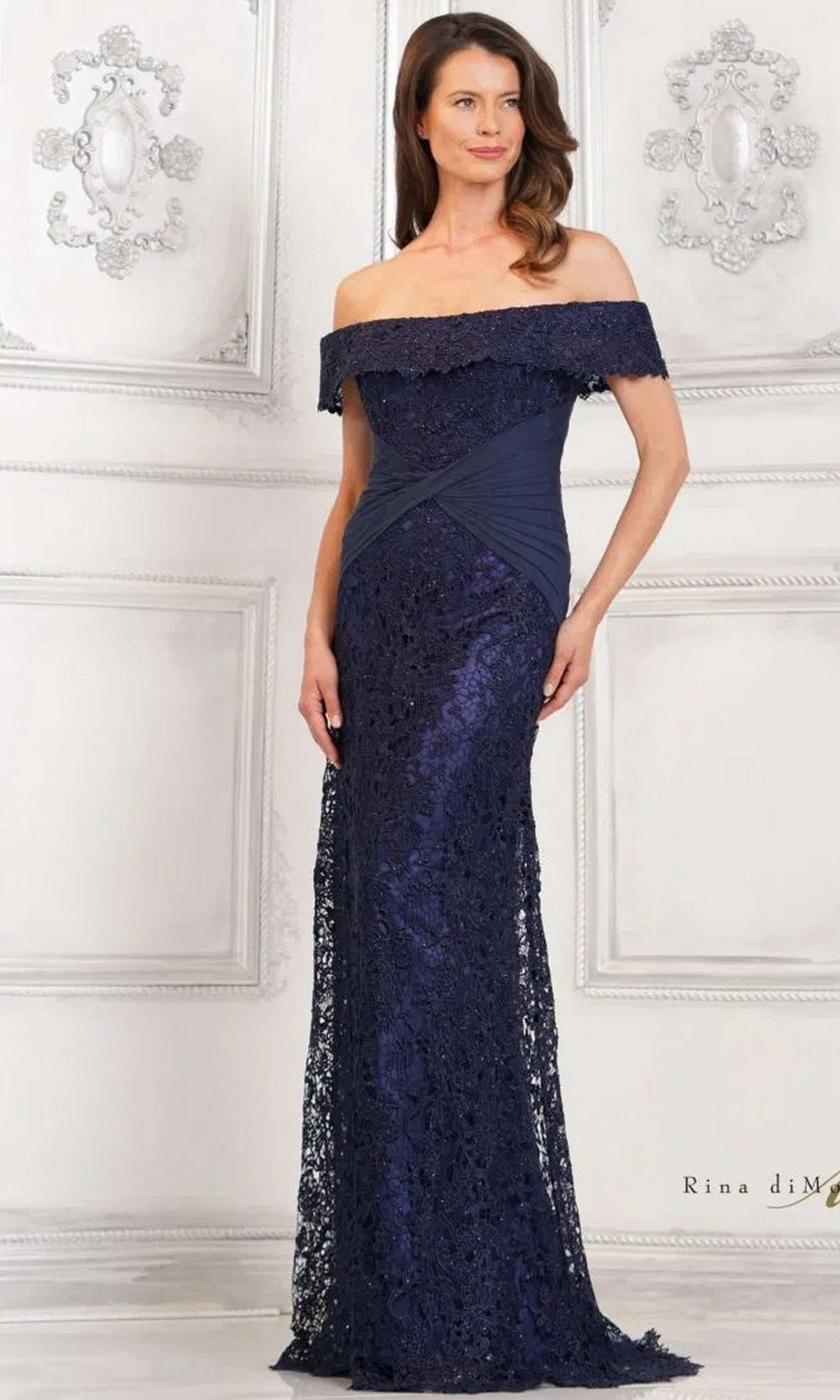 A woman stands gracefully against a white ornate wall, wearing the Rina Di Montella RD3129 long formal dress in navy.